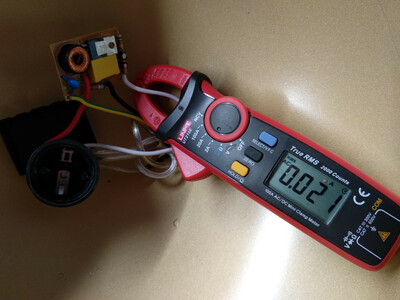 clamp multimeter reading 0.02 amps AC, connected to a small circuit board with a yellow capacitor, a coil, and a heat sinked IC visible